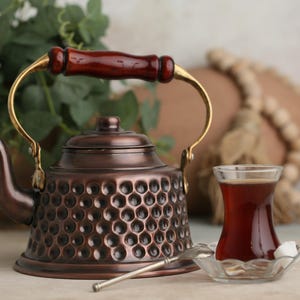 Peut inclure: Une théière en cuivre avec une finition martelée et une poignée en bois. La théière a un bec en laiton et un couvercle en laiton. Un verre de thé et un morceau de sucre sont sur une soucoupe à côté de la théière.