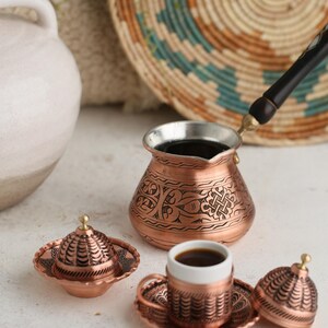 Op de afbeelding: Een koperen Turkse koffiepot met ingewikkelde ontwerpen, een bijpassend kopje en schoteltje, en een klein suikerpotje met deksel. De set staat op een wit oppervlak.