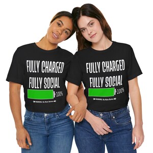 Puede incluir: Dos camisetas negras con texto blanco que dice "FULLY CHARGED FULLY SOCIAL" y un icono de batería verde que dice "100%". El texto "WARNING: No More Button" está debajo del icono de la batería.