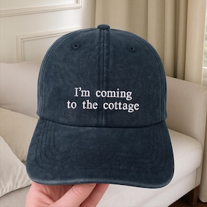 May include: A navy blue baseball cap with the embroidered text "I'm coming to the cottage" in white. The cap has a curved brim and a classic six-panel design. The hat is made of a soft, washed fabric.