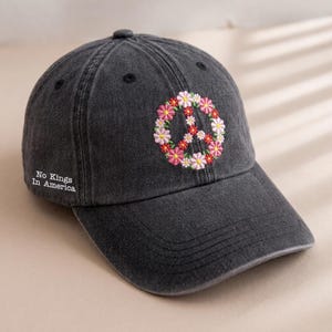 May include: A dark gray baseball cap with a curved brim. The cap features an embroidered peace sign made of pink, white, and red flowers. The text "No Kings In America" is embroidered on the side.