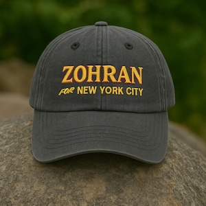 Könnte beinhalten: Graue Baseballkappe mit dem gelb gestickten Wort "ZOHRAN" und dem Text "FOR NEW YORK CITY". Die Kappe besteht aus weichem Material und hat einen gebogenen Schirm. Die Kappe liegt auf einem Stein.