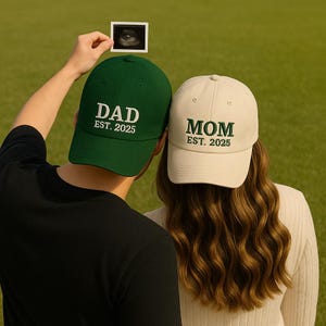Może przedstawiać: Dwie czapki bejsbolowe, jedna zielona z napisem "DAD EST. 2025" i druga kremowa z napisem "MOM EST. 2025". Pomiędzy nimi trzymane jest zdjęcie USG. Czapki są na zielonym tle, sugerując ogłoszenie.