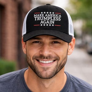 Op de afbeelding: Zwarte en witte truckerpet met de witte tekst "MAKE AMERICA TRUMPLESS AGAIN". De pet heeft een zwart voorpaneel en een witte mesh achterkant. De tekst staat boven en onder een rode lijn met witte sterren.