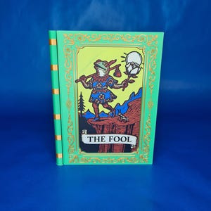 Può includere: Un mazzo di carte dei tarocchi verdi con la carta "Il Matto". La carta raffigura una figura simile a una rana in abiti colorati, che tiene un bastone e un fiore. La carta ha un bordo dorato e il testo "THE FOOL" in basso.
