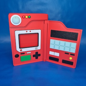Pode incluir: Console de jogos portátil vermelho com uma moldura de tela branca, botões verdes e pretos e um sinal de mais. O console aberto revela uma grade de botões azuis claros e pretos. O dispositivo está em um fundo azul.