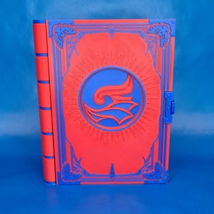 Op de afbeelding: Een rood en blauw decoratief boek met ingewikkelde details. De kaft heeft een blauw embleem in een cirkel, omgeven door sierlijke blauwe randen. De rug en sluiting zijn ook blauw.