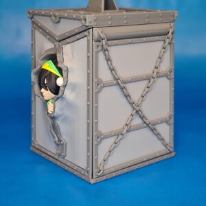 Op de afbeelding: Een grijze, doosvormige container met een handvat en kettingaccenten. Een stripfiguur met een groene en gele hoofdband kijkt uit een gat in de zijkant. De doos heeft een geklonken ontwerp.