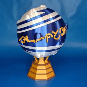 May include: A 3D-printed decorative sphere with blue and white stripes and gold lettering. The sphere sits on a gold-colored, multi-tiered base. The background is a solid blue.