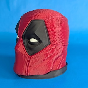 Può includere: Una maschera rossa di Deadpool con dettagli neri intorno agli occhi e alla bocca. La maschera ha una superficie strutturata e una base nera. Lo sfondo è blu.