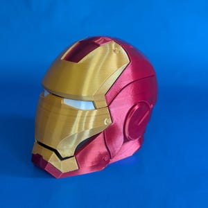 May include: A red and gold Iron Man helmet, a replica of the superhero's iconic headgear. The helmet features a gold faceplate and a red shell, with detailed design elements. The helmet is set against a blue background.