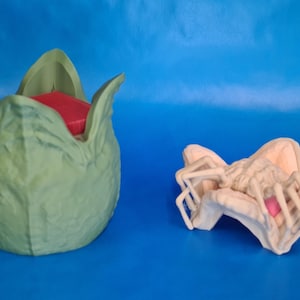 May include: Two fantasy sculptures against a blue backdrop. One is a large, pale green, plant-like form with a red interior. The other is a light beige, insect-like creature with a pink centre, possibly a prop or collectible.