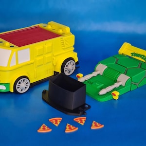 Op de afbeelding: Een gele en rode speelgoedbus met witte accenten, een groene voorkant en grijze wielen. Daarnaast staat een groen en geel voertuig met armen en een spoiler. Er is ook een zwarte container en pizzapunten.