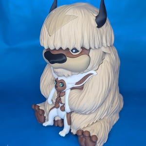 Op de afbeelding: Gedetailleerd beeldje van Appa, een bisonachtig wezen uit Avatar: The Last Airbender, in beige, bruin en zwart. Een kleiner figuur van Momo is aan Appa bevestigd. Het beeldje staat tegen een blauwe achtergrond.