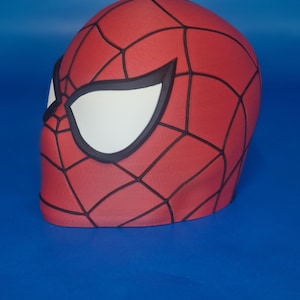 Puede incluir: Una máscara roja de Spider-Man con un patrón de telaraña negra y detalles blancos en los ojos. La máscara es un objeto tridimensional, probablemente para cosplay o disfraces. El fondo es azul liso.