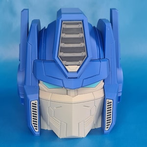 May include: A blue and gray Optimus Prime head, a toy or collectible, is displayed against a blue background. The head features detailed molding, including a faceplate, vents, and side panels. The design is reminiscent of the Transformers franchise.