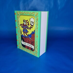 May include: A tarot card featuring a frog character holding a flower and a staff, with the text "THE FOOL". The card has a green border with gold accents and a yellow background. The book is upright with white pages.