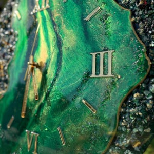 May include: A wall clock with a swirling emerald green and gold resin face featuring metallic Roman numerals and gold hands. The irregular, geode-inspired edge is encrusted with dark green and black crushed glass fragments and gold leaf accents.