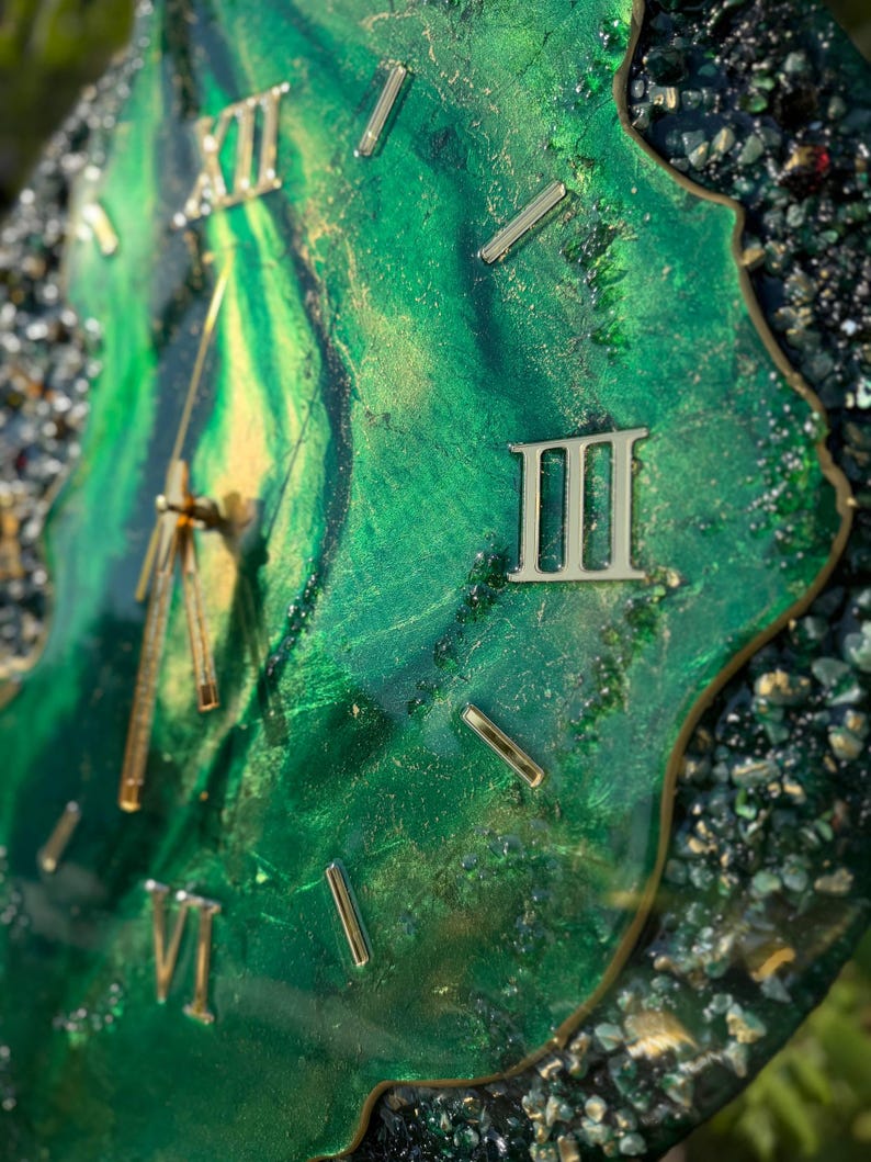 May include: A wall clock with a swirling emerald green and gold resin face featuring metallic Roman numerals and gold hands. The irregular, geode-inspired edge is encrusted with dark green and black crushed glass fragments and gold leaf accents.