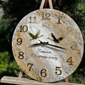 May include: Round wall clock with a gold-toned frame and numerals. The clock face features a marbled design with gold accents, two bird silhouettes, and text in a script font. The clock is displayed on a wooden easel.
