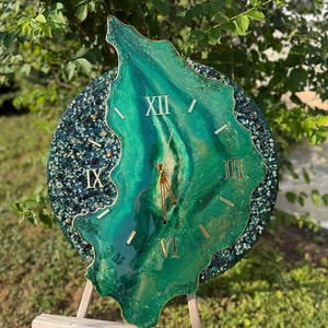 May include: A circular wall clock featuring a deep green resin center with swirling patterns and gold-colored Roman numerals. The outer edge is decorated with dark green and black crushed glass accents. Gold clock hands are positioned at the center.