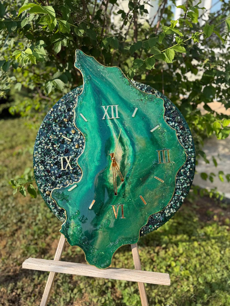 May include: A circular wall clock featuring a deep green resin center with swirling patterns and gold-colored Roman numerals. The outer edge is decorated with dark green and black crushed glass accents. Gold clock hands are positioned at the center.