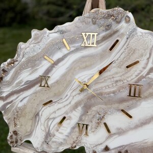 May include: A wall clock with an irregular, geode-inspired shape featuring swirling layers of white, cream and brown resin. The clock face includes gold-coloured Roman numerals at the XII, IX, VI and III positions, with gold hands.