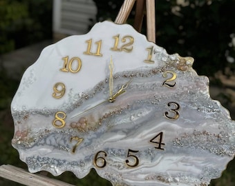 May include: A decorative wall clock with a unique, organic shape. The clock face features a white and gray marbled design with gold-colored numerals and clock hands. The clock is displayed on a wooden easel.