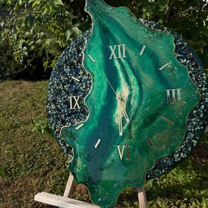 May include: A circular wall clock featuring a central organic shape in shades of emerald and forest green with gold accents. The clock face displays gold Roman numerals at the twelve, three, six, and nine o'clock positions with gold clock hands.