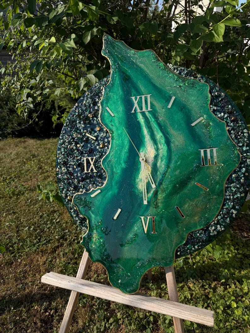 May include: A circular wall clock featuring a central organic shape in shades of emerald and forest green with gold accents. The clock face displays gold Roman numerals at the twelve, three, six, and nine o'clock positions with gold clock hands.