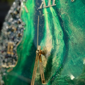 May include: A wall clock featuring a swirling emerald green and gold resin face with a glossy finish. The clock has gold-colored hands and Roman numerals, including a prominent XII at the top. The left edge is adorned with textured, dark crystal accents.