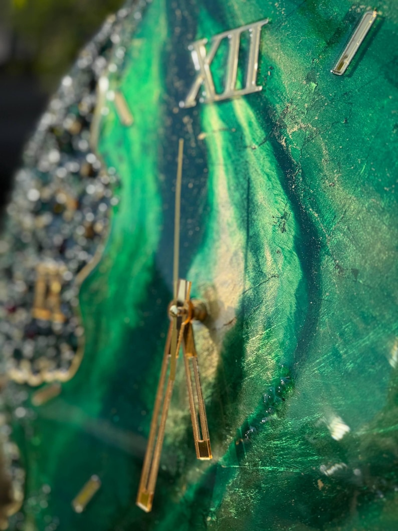 May include: A wall clock featuring a swirling emerald green and gold resin face with a glossy finish. The clock has gold-colored hands and Roman numerals, including a prominent XII at the top. The left edge is adorned with textured, dark crystal accents.