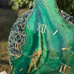 May include: A circular wall clock featuring a deep emerald green and gold marbled resin surface with a textured, crystalline border. The clock face displays gold-colored Roman numerals and thin gold hands, creating a layered, geode-inspired aesthetic.