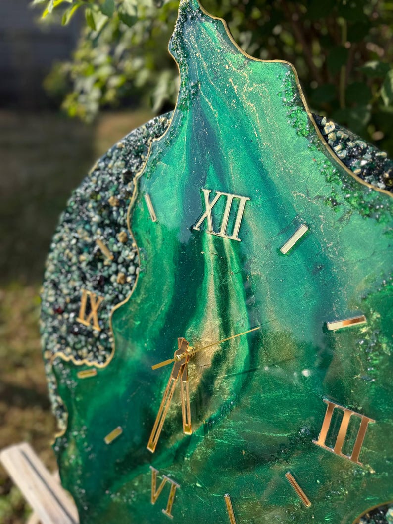 May include: A circular wall clock featuring a deep emerald green and gold marbled resin surface with a textured, crystalline border. The clock face displays gold-colored Roman numerals and thin gold hands, creating a layered, geode-inspired aesthetic.