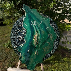 May include: A round wall clock with an irregular, organic-shaped center in shades of emerald green and gold. The clock face features gold Roman numerals at the XII, III, VI, and IX positions, with gold hands and a dark, textured, glittery border.