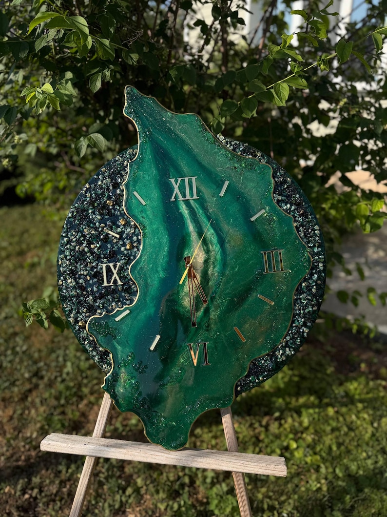 May include: A round wall clock with an irregular, organic-shaped center in shades of emerald green and gold. The clock face features gold Roman numerals at the XII, III, VI, and IX positions, with gold hands and a dark, textured, glittery border.
