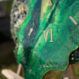 May include: A wall clock with an irregular, organic shape featuring a swirling emerald green and gold resin surface. The clock face includes metallic gold Roman numerals and slender gold hands, with textured, glittery dark edges.