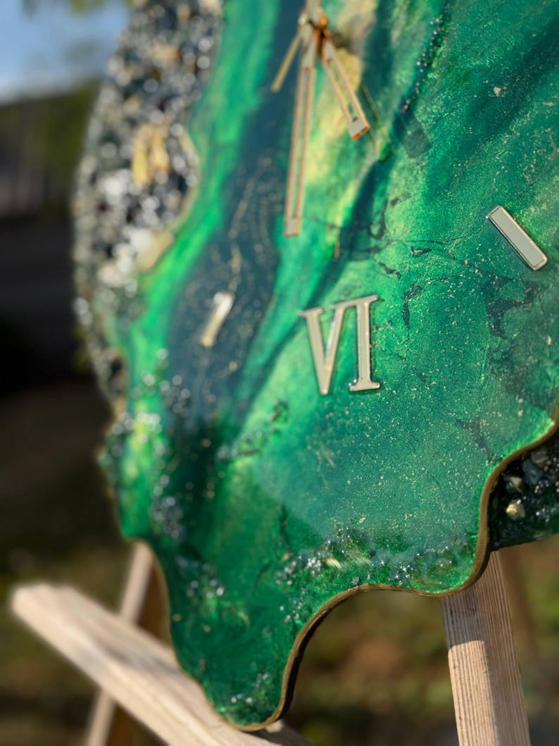 May include: A wall clock with an irregular, organic shape featuring a swirling emerald green and gold resin surface. The clock face includes metallic gold Roman numerals and slender gold hands, with textured, glittery dark edges.