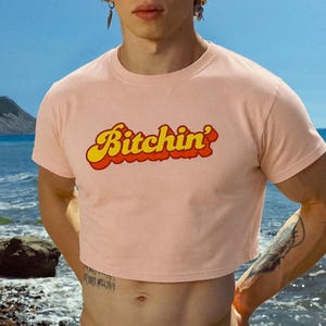 Könnte beinhalten: Ein hellrosa bauchfreies Kurzarm-Oberteil mit dem Wort Bitchin' auf der Brust, gedruckt in einer auffälligen, gelben Retro-Schrift mit dickem rotem Rand. Das Kleidungsstück hat einen einfachen Rundhalsausschnitt und einen geraden Saum an der Taille.