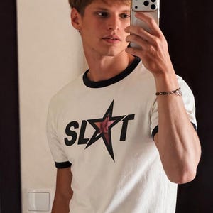 White t-shirt with black trim and a star graphic that reads "SLUT". The shirt is paired with blue jeans and a studded belt. The person in the image is holding a smartphone.