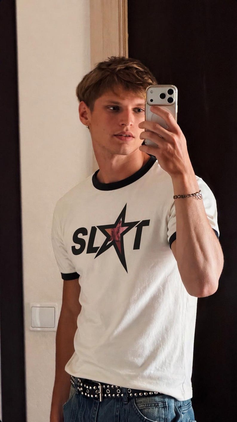 White t-shirt with black trim and a star graphic that reads "SLUT". The shirt is paired with blue jeans and a studded belt. The person in the image is holding a smartphone.