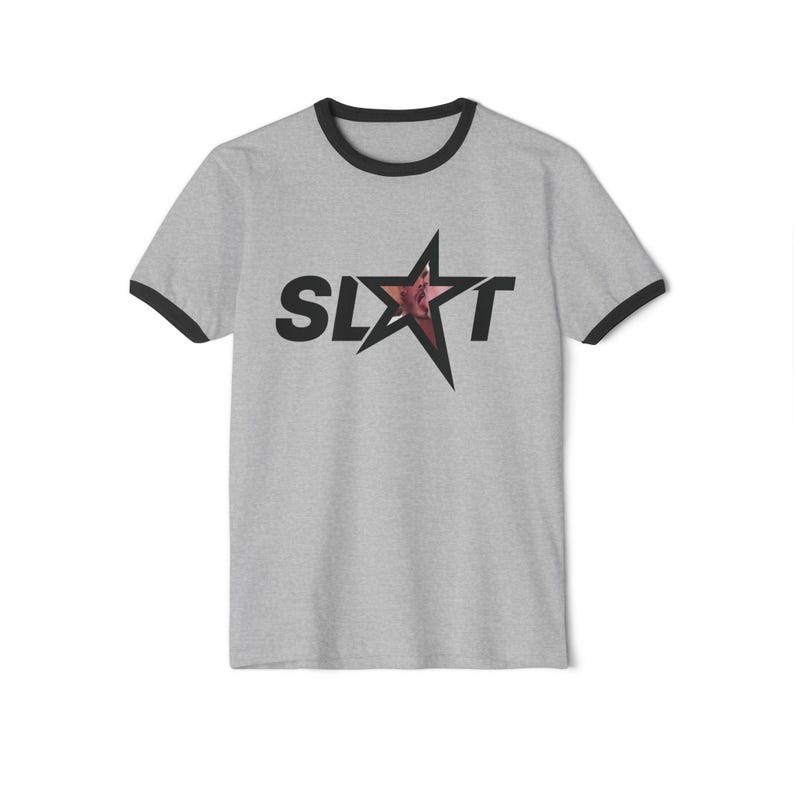 Puede incluir: Camiseta gris con ribetes negros en cuello y mangas. La camiseta presenta la palabra "SLXT" en letras negras con una estrella en el centro. La estrella tiene un dise&ntilde;o rosa y rojo. Cuello redondo y mangas cortas.