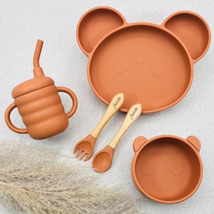 May include: A terracotta-coloured baby feeding set. It includes a bear-shaped plate and bowl, a sippy cup, and a fork and spoon. The wooden utensils are engraved with the name "Juliette". The set is made of silicone and wood.