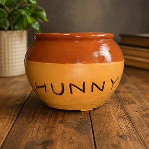 Può includere: Un vaso rotondo in ceramica bicolore, con la parte superiore marrone e la base gialla. La parola "HUNNY" è scritta in lettere nere sulla sezione gialla. Il vaso è appoggiato su una superficie di legno.