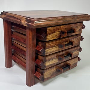 May include: A handcrafted wooden side table with a square top and four drawers. The table is made of reddish-brown and tan wood, with dark handles and decorative corner accents. The table is approximately 18 inches tall and 16 inches wide.