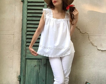 Puede incluir: Una blusa blanca de algodón con ribete de encaje y un pantalón blanco de talle alto con ribete de encaje. La blusa tiene un escote cuadrado y el pantalón tiene pierna ancha.