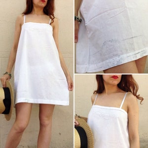 May include: A white cotton slip dress with thin straps and a simple embroidered design. The dress has a loose, flowing fit and a straight hemline.
