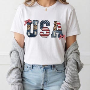 Peut inclure: T-shirt blanc avec le mot "USA" dans un design patriotique. Les lettres sont décorées de denim, d'imprimé léopard et du drapeau américain. Des nœuds rayés rouges et blancs accentuent les lettres. Le t-shirt est porté avec un short en jean bleu.