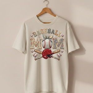 Peut inclure: T-shirt crème avec l'inscription "BASEBALL MOM" dans un motif floral. Le motif comprend une balle de baseball, un casque rouge et des battes croisées. Le t-shirt est suspendu à un cintre en bois.