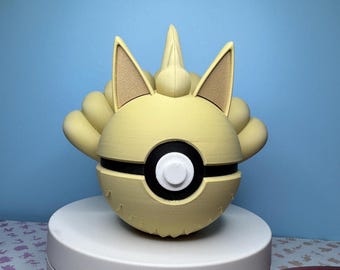 May include: A light yellow, spherical object with a black band and white center, resembling a stylized ball. It features tan-colored ear-like structures and a spiky top, set against a light blue background. The object appears to be a decorative item.